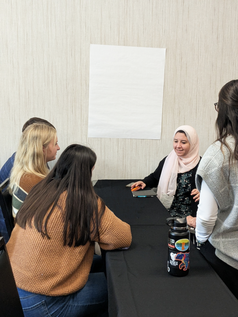 IPE Students discussing around a table. A sheet of large note paper hangs on the wall, ready for discussion.